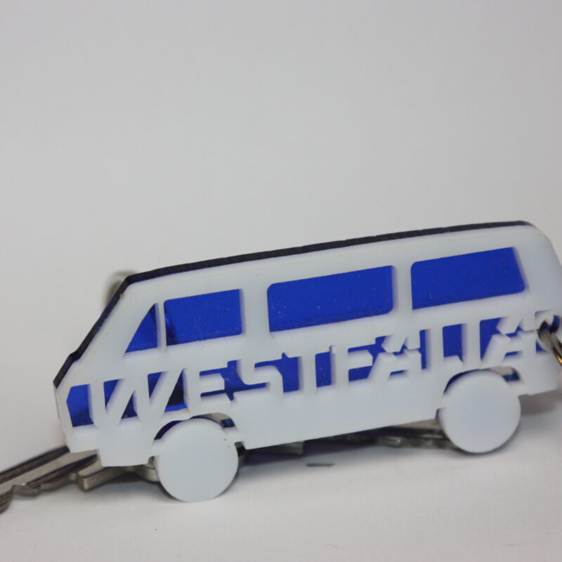 Ein wei&szlig;-blauer Westfalia-Schl&uuml;sselanh&auml;nger in Busform liegt neben einem Metallschl&uuml;ssel auf hellem Hintergrund.